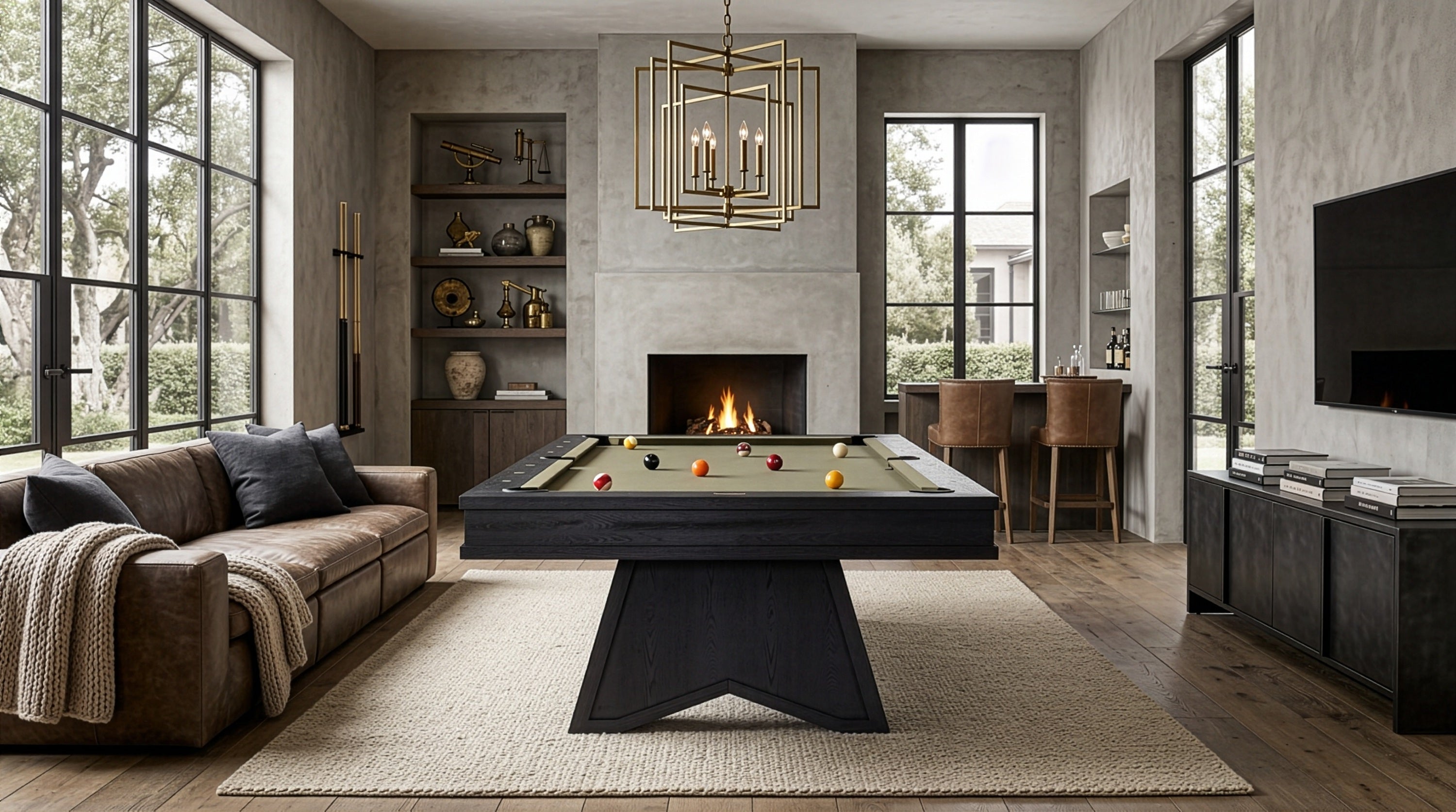 A modern living room featuring a dark wood pool table centered on a large beige rug, with a fireplace, large windows, a brown sofa, and a hanging brass chandelier.