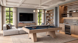 A modern living room featuring a light wood pool table with a dark blue felt surface, styled in a high-end contemporary space with a sectional sofa, built-in shelving, and a bar area.