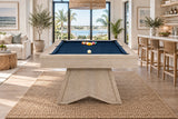 A light natural wood pool table with a dark blue felt surface, set in a bright, modern beach-themed living room with a large woven jute rug.