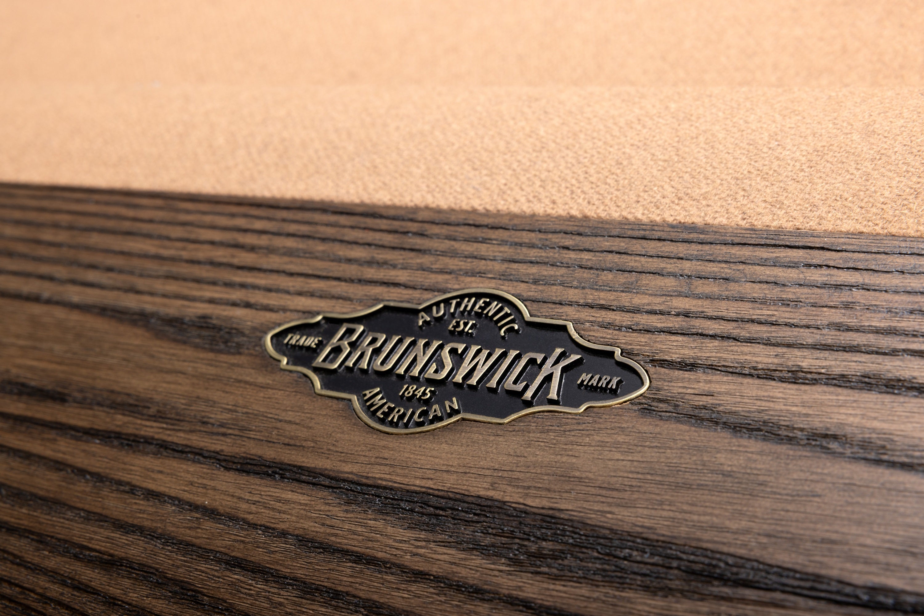 Close-up of a Brunswick hardwood pool table finish showing a dark brown wood grain surface with a brass logo plaque reading 'Authentic Brunswick 1965 American'.