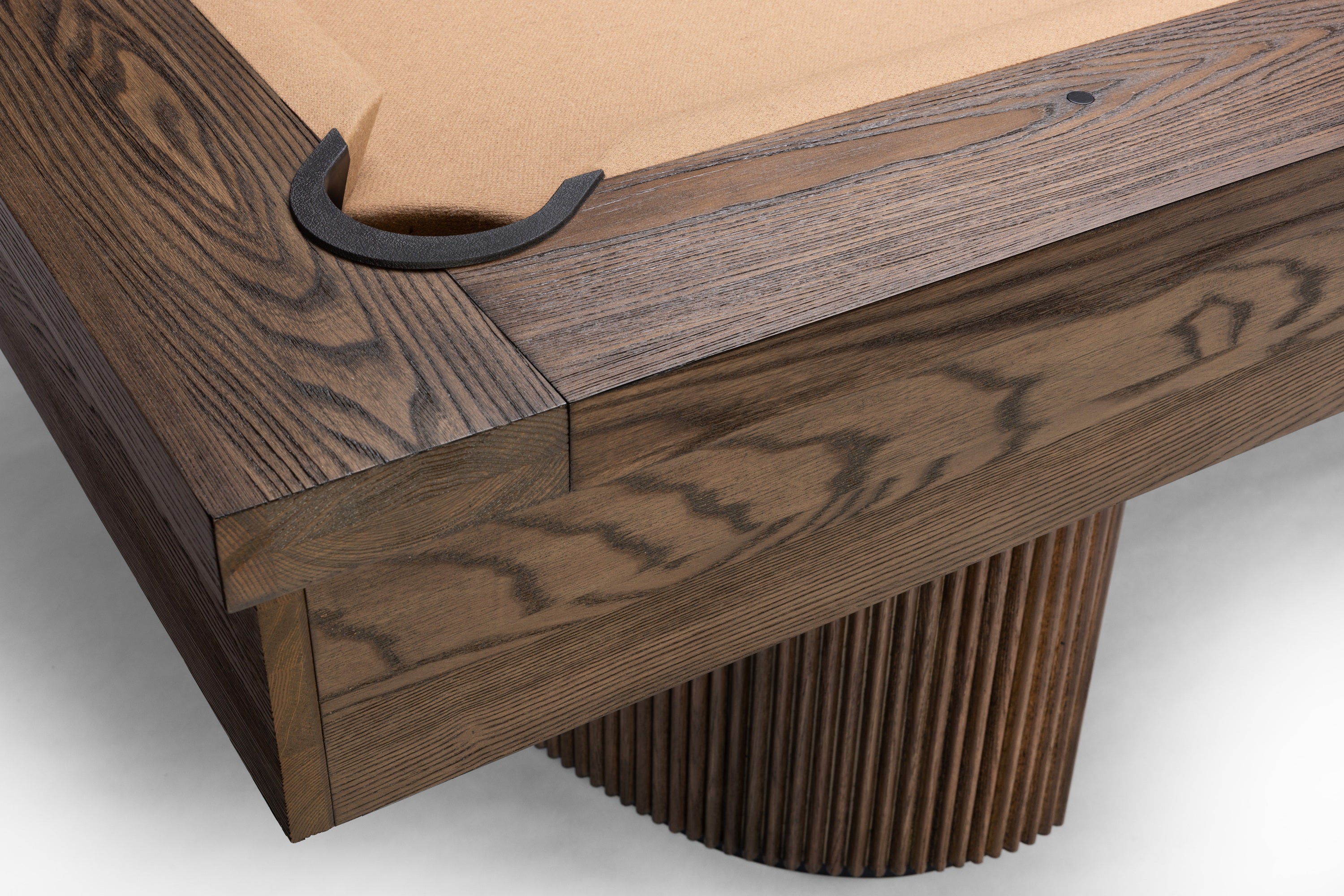 Close-up of a wooden pool table corner with a dark brown wood frame, tan felt surface, and a pleated cylindrical support leg on a light gray background.